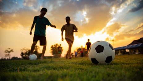 Silhouette action sport outdoors of a group of kids having fun playing soccer football for exercise in community rural area under the twilight sunset. Poor and poverty children in development country.